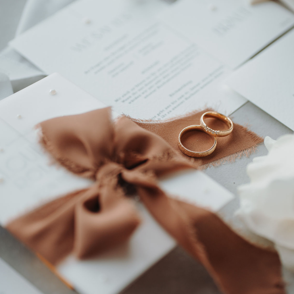 Flatlay Hochzeitspapeterie mit Fokus auf die bräunliche Schleife und die Hochzeitsringe